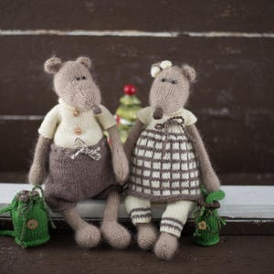 May include: Two knitted mouse toys, one brown with a white shirt and brown trousers, the other grey with a white and brown plaid dress, sitting on a white surface.