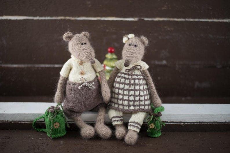 May include: Two knitted mouse toys, one brown with a white shirt and brown trousers, the other grey with a white and brown plaid dress, sitting on a white surface.