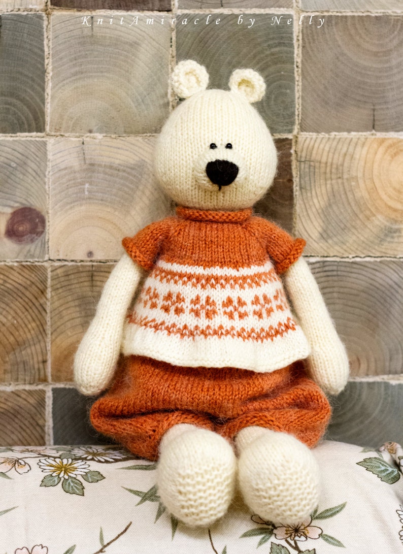 Puede incluir: Un oso de peluche blanco con un su&eacute;ter y pantalones marrones y blancos. El oso est&aacute; sentado en un coj&iacute;n con estampado floral.