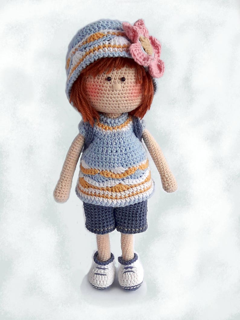 May include: A crocheted doll with red hair, wearing a blue and white striped hat with a pink flower, a blue and white striped shirt, blue shorts, and white shoes.