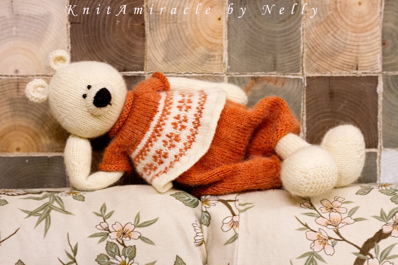 Puede incluir: Un oso de peluche blanco con un su&eacute;ter naranja y blanco con un patr&oacute;n geom&eacute;trico. El oso est&aacute; acostado sobre una tela de color crema con un patr&oacute;n floral.