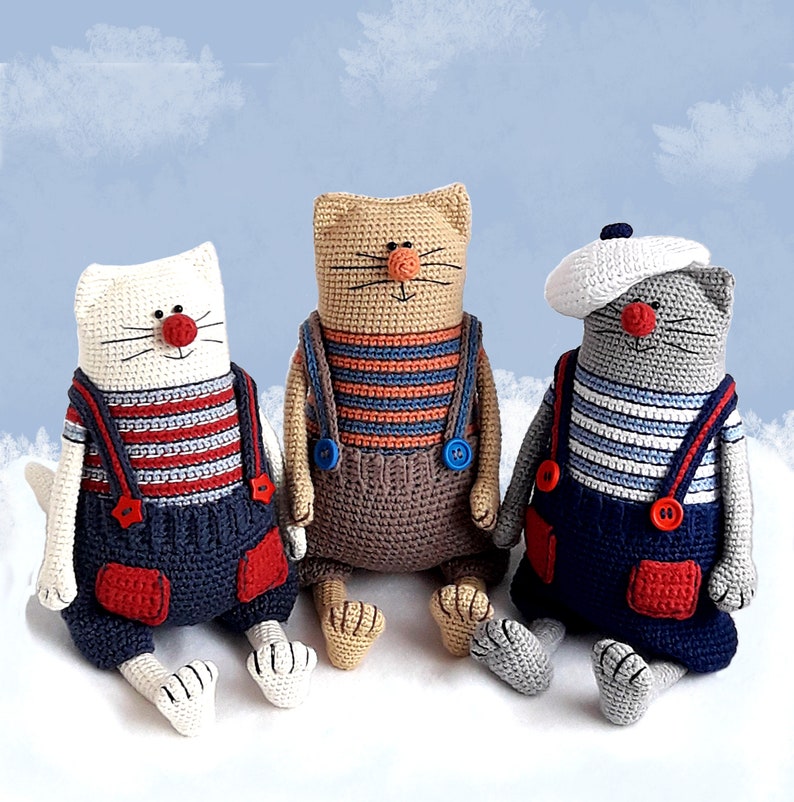 May include: Three crocheted cat toys with sailor outfits. The cat on the left is white with red and blue stripes, the middle cat is brown with orange and blue stripes, and the cat on the right is gray with white and blue stripes.