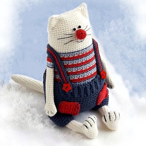 May include: A white crocheted cat wearing blue overalls with red accents and a red nose. The cat is sitting on a white fluffy surface.