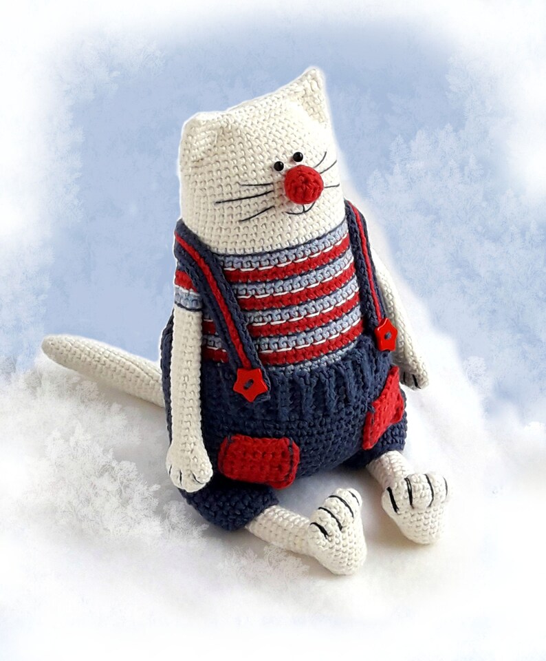 May include: A white crocheted cat wearing blue overalls with red accents and a red nose. The cat is sitting on a white fluffy surface.