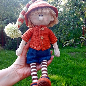 Puede incluir: Una mu&ntilde;eca de crochet con un gorro a rayas, un su&eacute;ter naranja, pantalones azules y calcetines a rayas. La mu&ntilde;eca sostiene un peque&ntilde;o pomp&oacute;n en su mano.