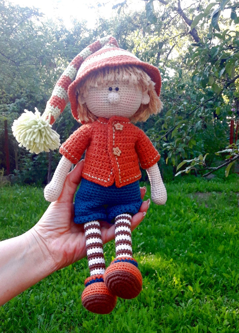 Puede incluir: Una mu&ntilde;eca de crochet con un gorro a rayas, un su&eacute;ter naranja, pantalones azules y calcetines a rayas. La mu&ntilde;eca sostiene un peque&ntilde;o pomp&oacute;n en su mano.