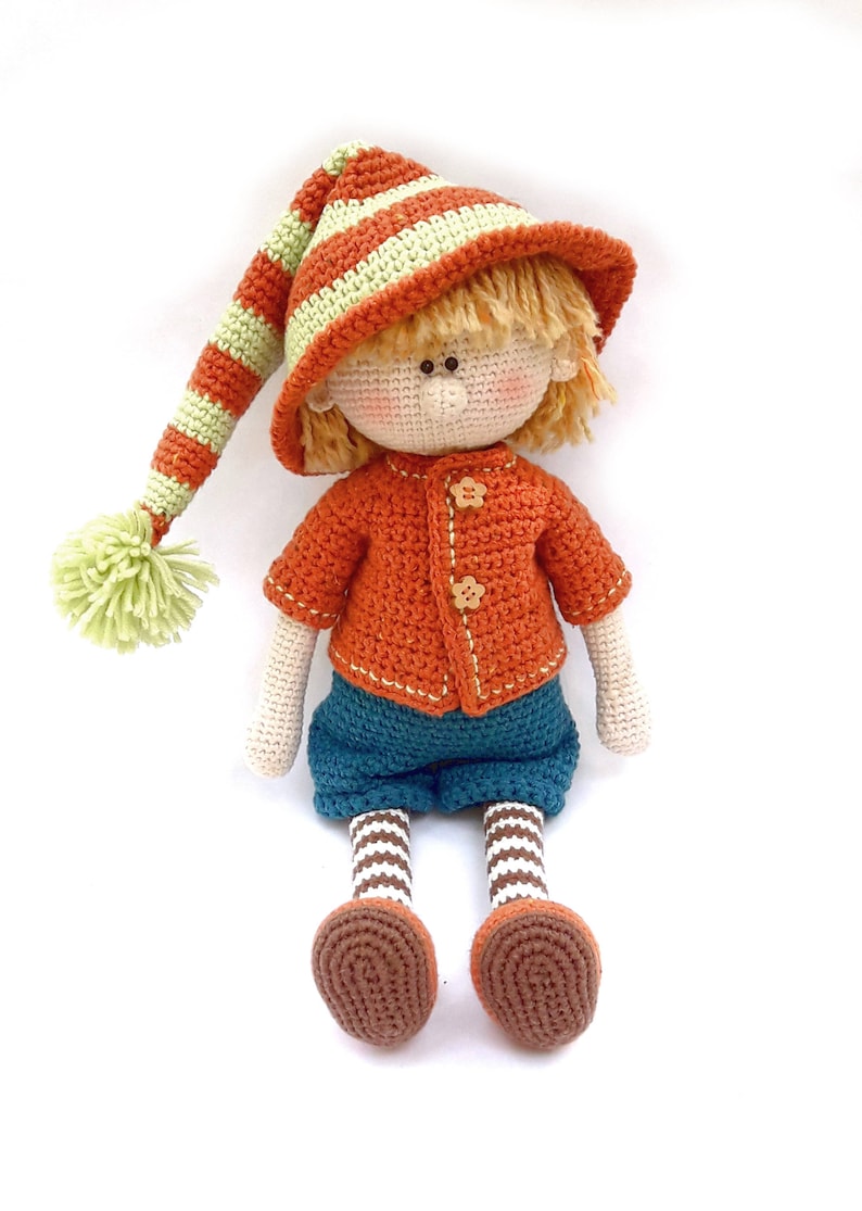 Puede incluir: Una mu&ntilde;eca de crochet con un sombrero a rayas, un su&eacute;ter naranja, pantalones azules y zapatos marrones. La mu&ntilde;eca tiene el pelo rubio y una cara sonriente.
