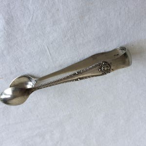 May include: Silver sugar tongs with an ornate design. The tongs are closed and have a slight curve.