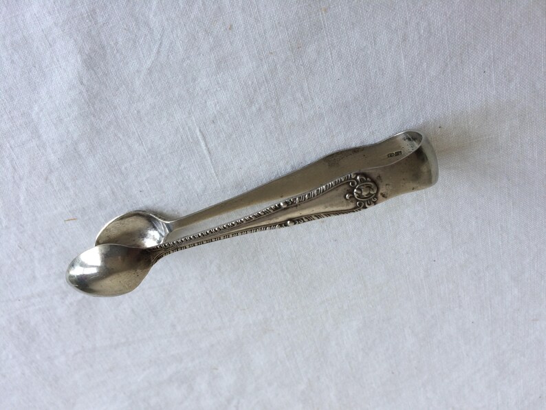 May include: Silver sugar tongs with an ornate design. The tongs are closed and have a slight curve.