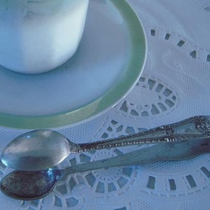 May include: Silver sugar tongs with a decorative design on a white lace tablecloth. The tongs are open and ready to be used.