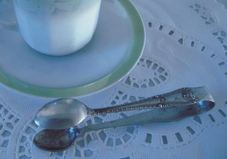 May include: Silver sugar tongs with a decorative design on a white lace tablecloth. The tongs are open and ready to be used.