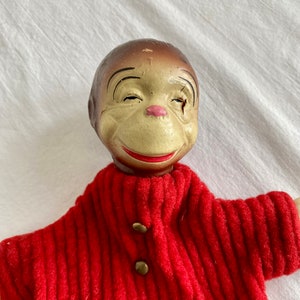 May include: A vintage hand puppet with a brown face, closed eyes, and a red sweater with gold buttons. The puppet is made of a hard material and has a painted face.
