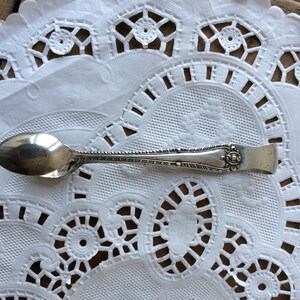 May include: A silver sugar tongs with an ornate design on the handle. The tongs are resting on a white doily.