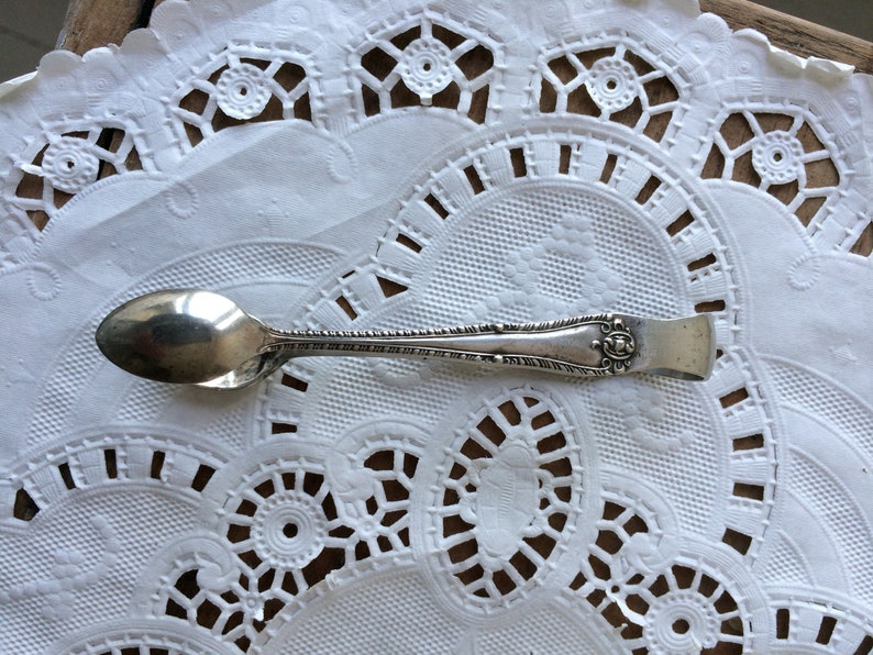 May include: A silver sugar tongs with an ornate design on the handle. The tongs are resting on a white doily.