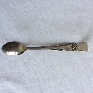 May include: Silver sugar tongs with an ornate design on the handle. The tongs have a rounded spoon-like end.