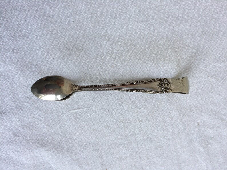May include: Silver sugar tongs with an ornate design on the handle. The tongs have a rounded spoon-like end.