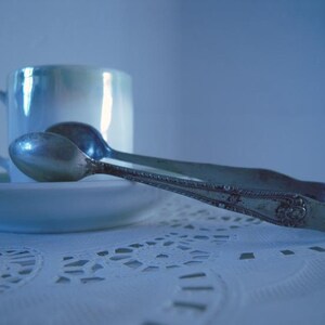 May include: A silver sugar tongs with an ornate design rests on a white lace tablecloth next to a white porcelain teacup.
