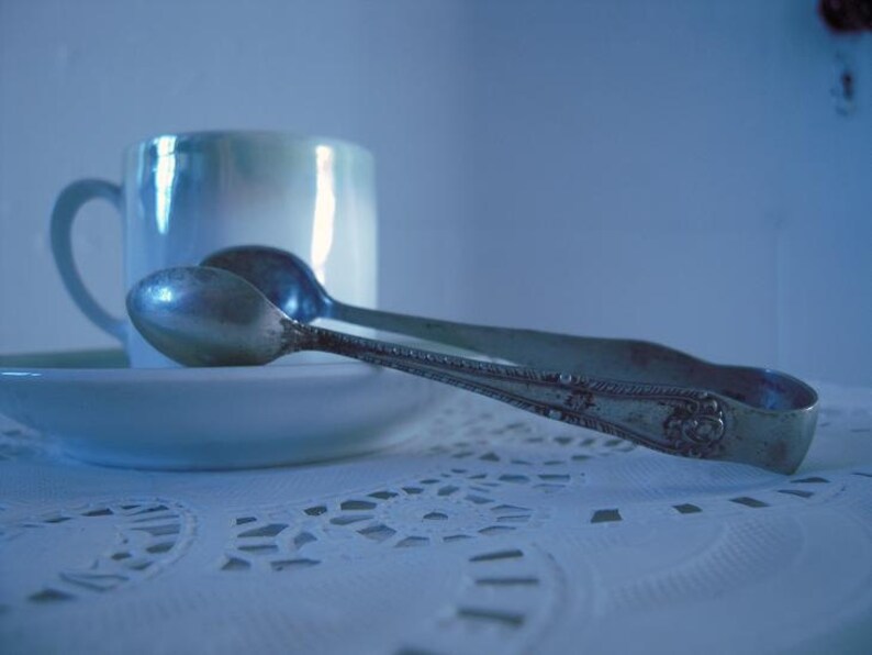 May include: A silver sugar tongs with an ornate design rests on a white lace tablecloth next to a white porcelain teacup.
