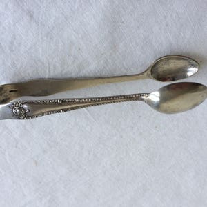May include: Silver sugar tongs with an ornate design. The tongs are made of two spoons joined together at the handle.