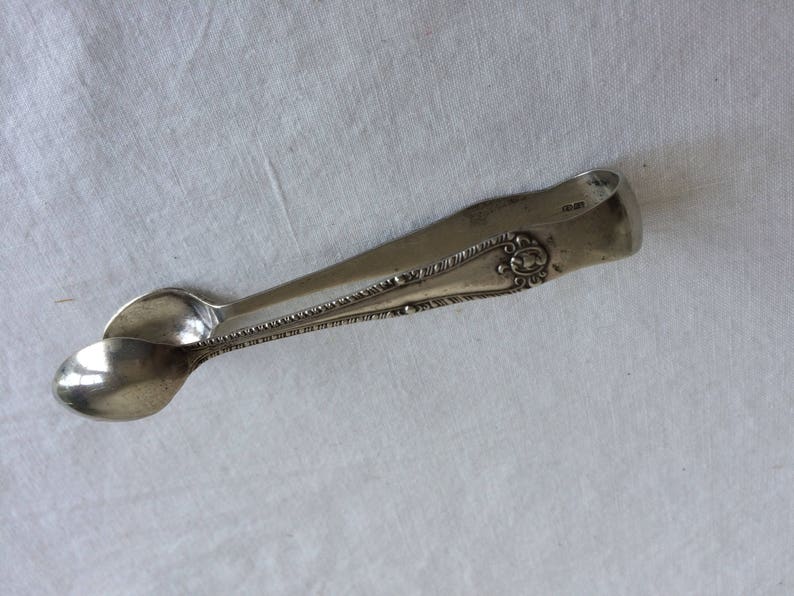 May include: Silver sugar tongs with an ornate design and the inscription "1883" on the handle.