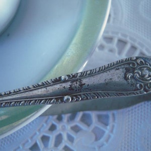 May include: Silver sugar tongs with an ornate design. The tongs are resting on a white tablecloth with a lace pattern.