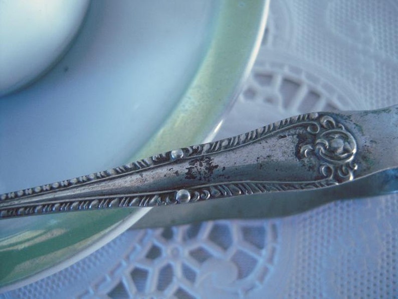 May include: Silver sugar tongs with an ornate design. The tongs are resting on a white tablecloth with a lace pattern.