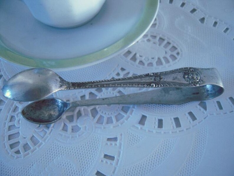 May include: Silver sugar tongs with an ornate design. The tongs are on a white lace tablecloth.