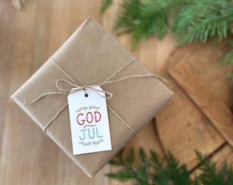 May include: A wrapped gift in brown paper, tied with twine, and a tag that reads "GOD JUL" in red and blue. The gift is on a wooden surface, with greenery and a straw star in the background.