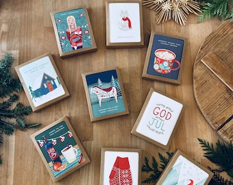 May include: A collection of Christmas greeting cards in brown boxes, featuring various festive illustrations. Designs include a woman in traditional clothing, a fox, a mug of hot cocoa, a house in the snow, and a skier. The cards are arranged on a wooden surface.