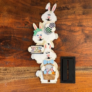 May include: Three white bunny figurines stacked on top of each other. The top bunny is holding a purple and green Easter egg. The middle bunny is holding a sign that says "Happy Easter". The bottom bunny is holding a basket filled with colorful Easter eggs.