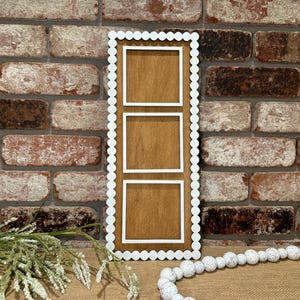 May include: A wooden picture frame with three square openings. The frame has a white scalloped border and is mounted on a brick wall.