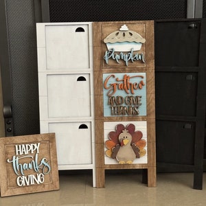 May include: A wooden tiered shelf with three white shelves and a brown shelf with Thanksgiving-themed decorations. The decorations include a pumpkin pie, a turkey, and a sign that says "Gather and Give Thanks". There is also a separate wooden sign that says "Happy Thanks Giving".