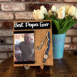 May include: A wooden picture frame with a fishing rod design. The frame has a black and white photo of a man and a young girl. The frame has a black sign that says "Best Papa Ever".