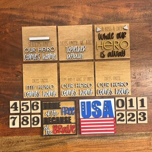 May include: A collection of wooden signs and number tiles. The signs feature phrases like "Our Hero Comes Home" and "Home of the Free Because of the Brave." The numbers are in black and white. The USA sign has a red, white, and blue color scheme.