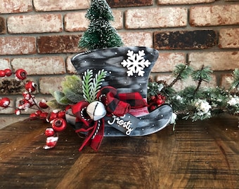 May include: A decorative black and white painted boot with a snowflake, a small Christmas tree, and a red and black plaid bow. The boot has the word "frosty" on a tag and is decorated with red berries and greenery.