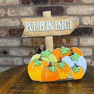 Puede incluir: Un letrero de madera con las palabras "PUMPKIN PATCH" en letras blancas. El letrero tiene forma de flecha y está montado sobre un poste de madera. Debajo, varias calabazas de madera coloridas en tonos naranja, amarillo y azul.