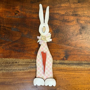 May include: A white wooden bunny with a pink and white plaid dress, a red heart, and a pink and white flower with a button in the center. The bunny is standing on a wooden surface.