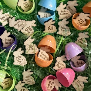 May include: A collection of plastic Easter eggs filled with various prizes, including a new book, a family craft, a game night, a pizza night, and a sweet treat. The eggs are surrounded by green grass and shredded paper.