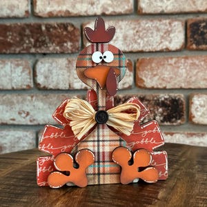 May include: A wooden turkey figurine with a plaid pattern, a brown bow, and the word "gobble" written on its wings. The turkey has large, round eyes and a red beak.