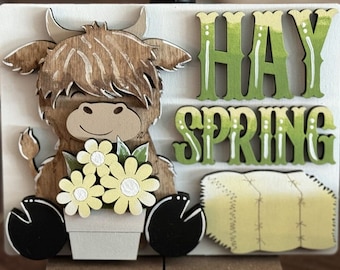 May include: A wooden sign with a brown and white highland cow holding a bouquet of yellow flowers. The sign says "Hay Spring" in green letters with white accents and a yellow hay bale.