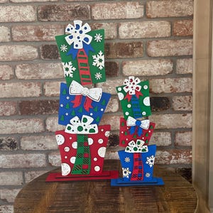 May include: A stack of colorful, decorative gift boxes in red, blue, and green, with white bows and snowflake accents. The boxes are arranged on a wooden surface against a brick wall, creating a festive holiday display.