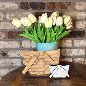 May include: A display featuring a bouquet of cream-colored tulips in a blue ceramic pot, set above a wooden picnic basket and a miniature version. The basket and miniature are made of light brown wood. The background is a brick wall.