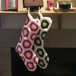 Könnte beinhalten: Zwei gehäkelte Weihnachtsstrümpfe hängen an einem Kaminsims. Ein Strumpf ist rot und weiß mit einem geometrischen Muster. Der andere Strumpf ist grün und weiß mit einem geometrischen Muster.