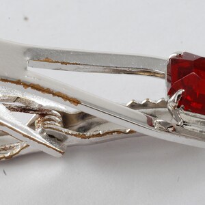 May include: A silver tie clip with a red, faceted gemstone. The clip has a spring mechanism and is slightly worn.