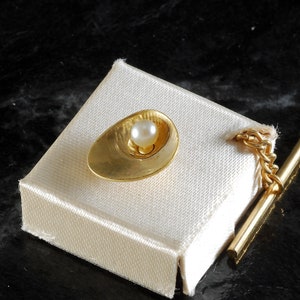 May include: A gold tie tack with a small pearl in the center. The tie tack is on a white background.