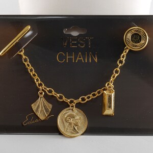 May include: Gold-toned vest chain with a bar pin, chain, and decorative charms. Charms include a fan shape, a coin with a profile, and a rectangular shape. The chain is displayed on a black card with the text "VEST CHAIN" and the signature "Eduardo".