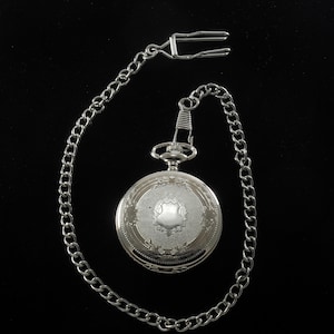 Puede incluir: Reloj de bolsillo plateado con un diseño grabado decorativo y una cadena a juego. El reloj tiene una forma redonda clásica y un acabado plateado. La cadena incluye un cierre para sujetarlo a la ropa.