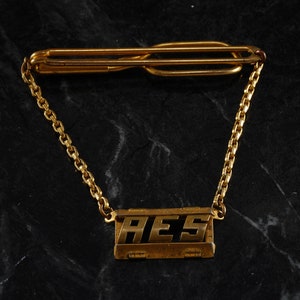 May include: A gold tie bar with a chain and a rectangular tag that says "AES".