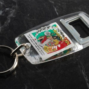 May include: A clear plastic bottle opener keychain with a cartoon image of a woman in a red and green costume. The image has the text "Never mind the safety faster! Just swallow the big one!"