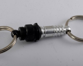 May include: A silver and black key chain with a retractable cord. The cord is attached to a small, black, cylindrical object with a silver ring on one end and a silver ring on the other end.